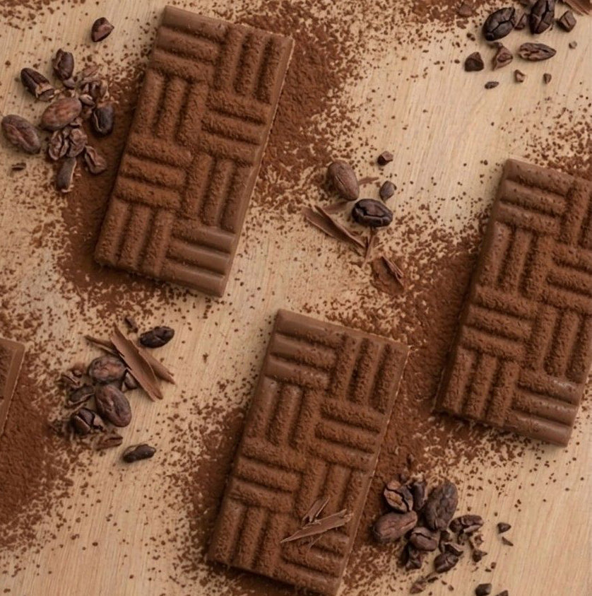Five rectangular chocolate wafers with a basketweave pattern, arranged on a light wooden surface with scattered cocoa powder, coffee beans, and cinnamon sticks.